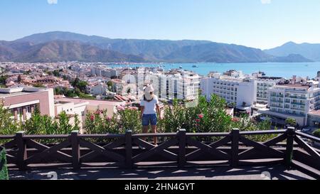 Donna in cappello cowboy si trova dietro la recinzione e guardando in distanza sul ponte di osservazione sopra la città di villeggiatura Marmaris in Turchia. Città turistica sul mare Foto Stock