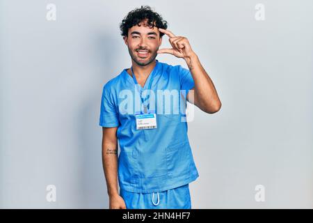 Giovane uomo ispanico che indossa l'uniforme blu maschio infermiera sorridente e fiducioso gesturing con mano facendo piccolo segno di dimensione con le dita guardando e la fotocamera Foto Stock