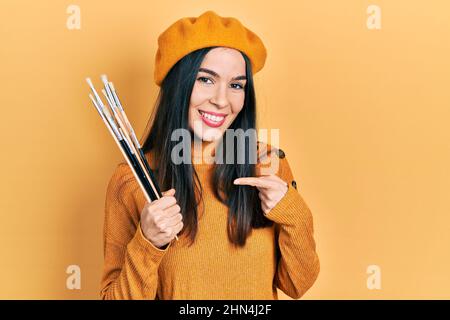 Giovane donna brunette che indossa artista look con beretto che tiene pennelli sorridenti puntamento felice con mano e dito Foto Stock