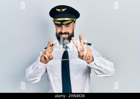 Giovane uomo ispanico che indossa l'uniforme di pilota di aeroplano sorridente guardando la macchina fotografica mostrando le dita facendo segno di vittoria. Numero due. Foto Stock
