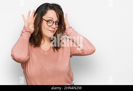 Giovane donna più misura indossando abiti casual e occhiali cercando di sentire entrambe le mani sul gesto orecchio, curioso per pettegolezzi. Problema uditivo, sordi Foto Stock