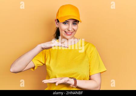 Giovane donna ispanica che indossa la divisa di consegna e cappuccio gesturing con le mani che mostrano grande e grande segno di misura, simbolo di misura. Sorridente guardando la camma Foto Stock