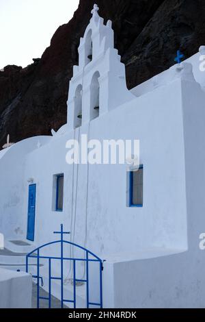 Vista di una bianca chiesa greco-ortodossa con una bandiera greca e il paesaggio vulcanico della famosa spiaggia rossa sullo sfondo Foto Stock