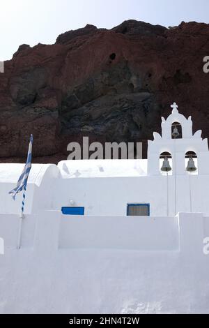 Vista di una bianca chiesa greco-ortodossa con una bandiera greca e il paesaggio vulcanico della famosa spiaggia rossa sullo sfondo Foto Stock