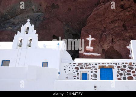 Vista di una bianca chiesa greco-ortodossa con una bandiera greca e il paesaggio vulcanico della famosa spiaggia rossa sullo sfondo Foto Stock