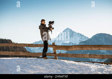 Uomo videografo che realizza riprese utilizzando una fotocamera montata su un dispositivo stabilizzatore gimbal. Neve e montagne sullo sfondo. Foto Stock