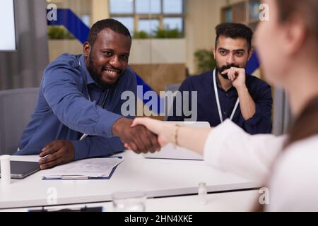 Felice giovane imprenditore afroamericano che scuote la mano di donna esperta in camice da laboratorio su scrivanie con documenti medici o finanziari Foto Stock