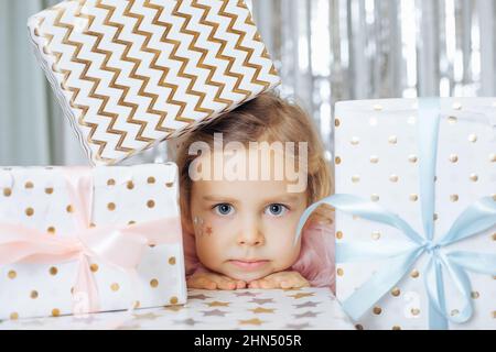 Bambina con capelli corti ricci e stelle sulla faccia in abito rosa cucciolo guardando fuori da mucchio di scatole regalo avvolte. Foto Stock