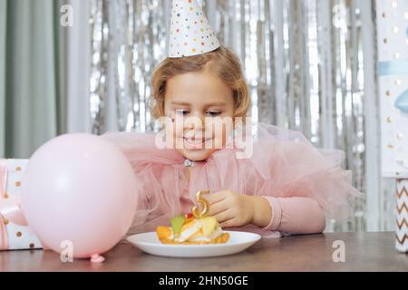 Bambina con capelli ricci e stelle sulla faccia in rosa vestito da cucciolo e cappuccio guardando la torta di frutta con la candela numero cinque. Foto Stock