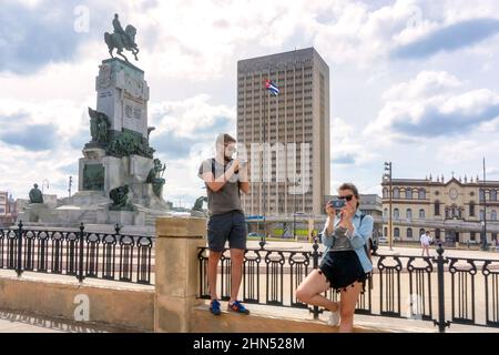 Turisti stranieri che scattano foto al Parco Antonio Maceo, dove è possibile ammirare una scultura. L'ospedale Hermanos Ameijeiras è visto sullo sfondo. Foto Stock