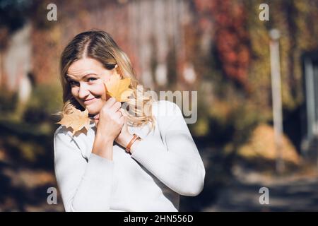 Donna piuttosto sorridente con capelli lunghi e ondulati in maglione roll-neck tenere le foglie di acero giallo vicino al viso con le braccia incrociate. Foto Stock