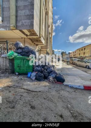 Cestino pieno di rifiuti nella strada sul marciapiede vicino al centro ElQods a Cheraga, Algeri. Persone che camminano e cas parcheggiato Foto Stock