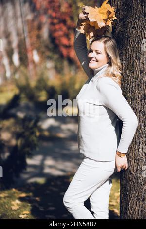 Donna sorridente beauteous di mezza età con i capelli lunghi e ondulati leali che alzano la mano con il bouquet delle foglie cadute che si appoggiano sull'albero. Foto Stock