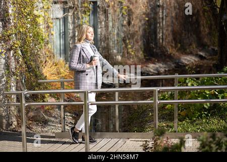 Attraente donna di mezza età in giacca a scacchi e maglione grigio con collo a rollino si distingue per il caldo sole autunnale. Foto Stock