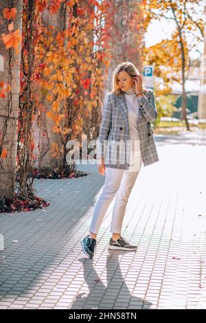 Graziosa donna di mezza età in giacca a scacchi, pullover grigio con girocollo e jeans bianchi posa i capelli titivanti vicino all'edificio. Foto Stock