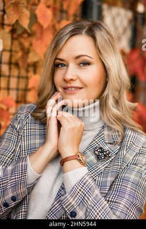 Donna sorridente di mezza età con capelli lunghi e leali in giacca a scacchi posa con la palma ripiegata vicino al viso su sfondo di edera. Foto Stock