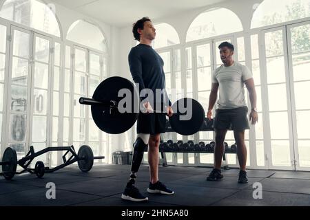 Ampio colpo di disabili atleta sollevamento pesi con il suo allenatore in palestra. Uomo con gamba protesica che viene allenato dal suo istruttore Foto Stock