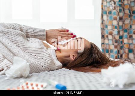 Una giovane donna caucasica si trova sul letto, soffia il naso in un fazzoletto. Vista laterale. Concetto di infezione virale e quarantena. Foto Stock