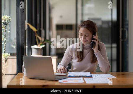 Ritratto della giovane donna asiatica seduta alla sua scrivania che lavora con calcolatrice e documenti mentre chiama il telefono cellulare in un ufficio moderno. Foto Stock