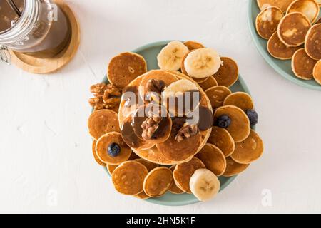 vista dall'alto di molte piccole frittelle e frittelle spesse con pasta di cioccolato su un piatto di ceramica. fette di banana, noci. colazione deliziosa Foto Stock
