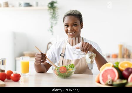 Sorridente donna afroamericana millenaria in grembiule fare l'insalata a tavola Foto Stock