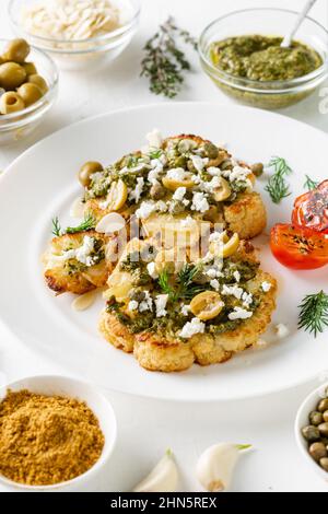 Bistecca di cavolfiore con spezie, salsa di chimichurri, fiocchi di mandorle, olive, pomodori ciliegini fritti e capperi su un piatto bianco. Cibo vegetariano. Foto Stock