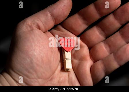 Cuore su un chiodo di vestiario nel palmo di un uomo su uno sfondo scuro. Il concetto di amore, le dichiarazioni di amore, il giorno di San Valentino. Foto Stock