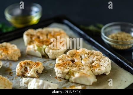 Cucina con bistecca di cavolfiore. Il cavolfiore crudo cosparso di spezie si trova su una teglia da forno. Olio d'oliva, erbe, varie spezie nelle vicinanze. Sfondo scuro. Foto Stock