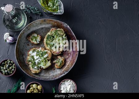 Bistecca di cavolfiore con spezie, salsa di chimichurri, fiocchi di mandorle. Capperi, olive, erbe, fianco a fianco. Sfondo scuro. Cibo vegetariano. CopySpace. Stagno Foto Stock