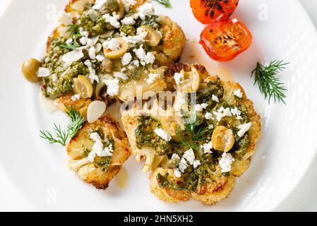 Bistecca di cavolfiore con spezie, salsa di chimichurri, fiocchi di mandorle, olive, pomodori ciliegini fritti e capperi su un piatto bianco. Cibo vegetariano. Foto Stock