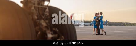 Tiro a tutta lunghezza di pilota maschile in posa per foto insieme a due hostess in uniforme blu, in piedi in un terminal aeroportuale al tramonto Foto Stock
