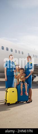 Scatto a tutta lunghezza di belle stewardesses in piedi vicino a due bambini seduti sul loro bagaglio di fronte al grande aereo e sorridenti alla macchina fotografica Foto Stock