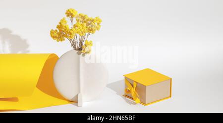 Fiori gialli immortelle eterni in vaso bianco in ceramica minimalista e scatola regalo gialla con fiocco a nastro su sfondo giallo e bianco. Greetin Foto Stock