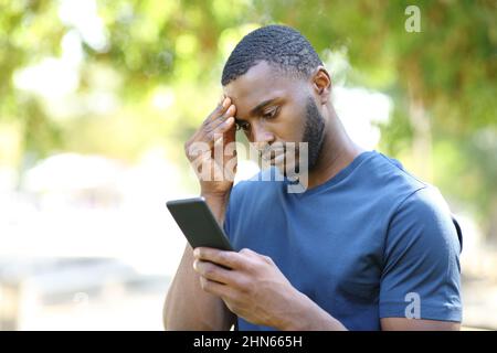 Uomo preoccupato con pelle nera che controlla il telefono cellulare in piedi in un parco Foto Stock