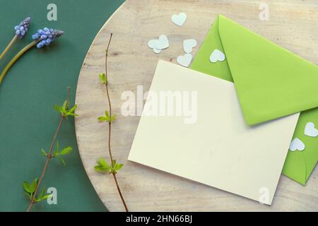 Sfondo primaverile in tonalità di verde. Busta di carta con bigliettino bianco e cuore confetti in pietra beige su tavola verde scuro. Foto Stock