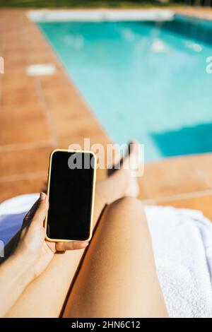 Donna irriconoscibile usando uno smartphone mentre si prende il sole in piscina Foto Stock