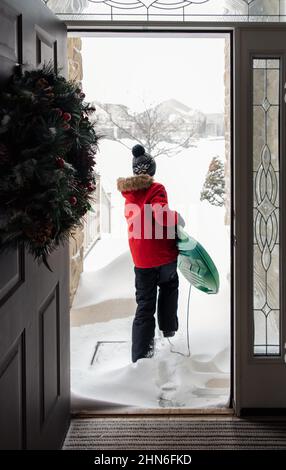 Ragazzo in cappotto rosso che esce dalla porta con slitta il giorno d'inverno. Foto Stock