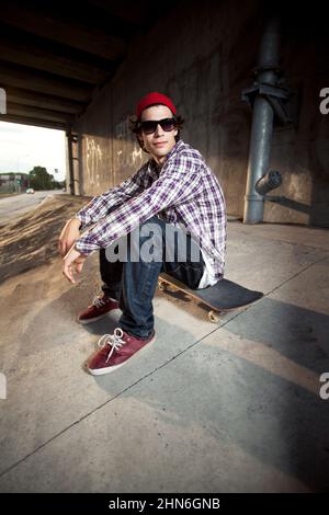 Skateboarder sotto il cavalcavia seduto con occhiali da sole Foto Stock