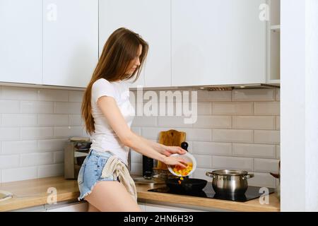 Giovane donna in piedi di profilo vicino alla stufa elettrica e mette le verdure tritate in una padella. Cute donna preparare salsa di verdure per la pasta a h Foto Stock