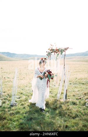 Sposa in una corona con un bouquet cammina oltre l'arco di nozze Foto Stock