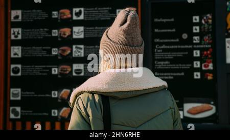 La donna torna alla macchina fotografica in un'area urbana di Street food sceglie il cibo da un menu. Street food festival. Giornata di sole in inverno o in autunno Foto Stock