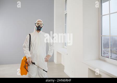 Sterminatore di controllo di peste in una maschera e vestito in piedi nella casa che tiene il suo irroratore insetticida Foto Stock