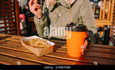 Donna che mangia gustose patatine fritte e beve te' o caffe' nella caffetteria all'aperto. Donna in un cortile urbano fuori strada cibo. Foto da primo piano Foto Stock