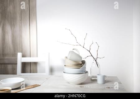 Una piramide di ciotole bianche, piatti e tazze di diverse dimensioni e colori su un tavolo beige in cucina. Un ramo di tiglio non soffiato in un vaso di vetro. SCA Foto Stock