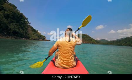 Giovane uomo con occhiali da sole e cappello ranghi rosa canoe di plastica lungo il mare contro verdi isole collinari con giungle selvatiche. Viaggiare in paesi tropicali. Il ragazzo forte sta navigando in kayak nell'oceano, vista posteriore. Foto Stock