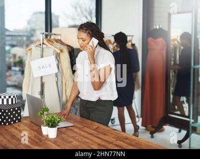 E come posso aiutarla oggi. Shot di un giovane imprenditore che risponde al suo cellulare e guarda il suo laptop nel suo negozio. Foto Stock