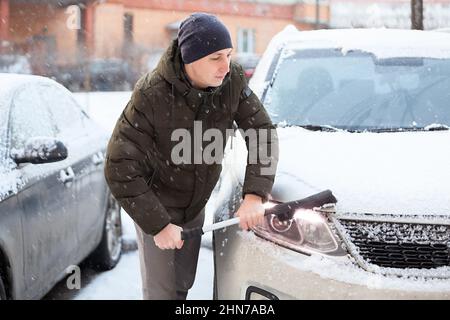 Un uomo caucasico adulto con spazzola di neve in mano che puliva la sua auto suv nella stagione invernale, facendo scorrere la neve dal faro Foto Stock