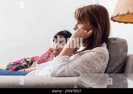 Giovane donna con sindrome di Down sdraiata sul divano guardando la macchina fotografica con la mamma, condividere con la famiglia, parlare e guardare la TV, stile di vita concetto w Foto Stock