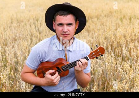 Stella della gioventù. Giovane bel caucasico uomo giocare ukulele fuori. Il bel ragazzo hipster suona la chitarra ukulele su sfondo naturale. Maschio in un cowboy Foto Stock