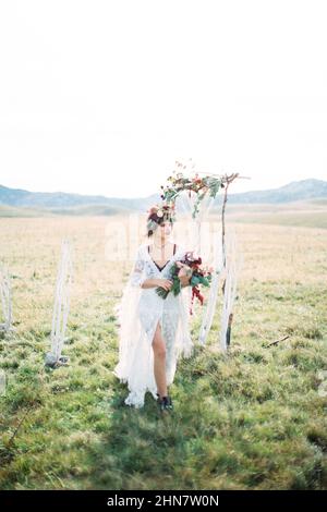Sposa in una corona cammina davanti ad un arco di nozze su un prato verde Foto Stock